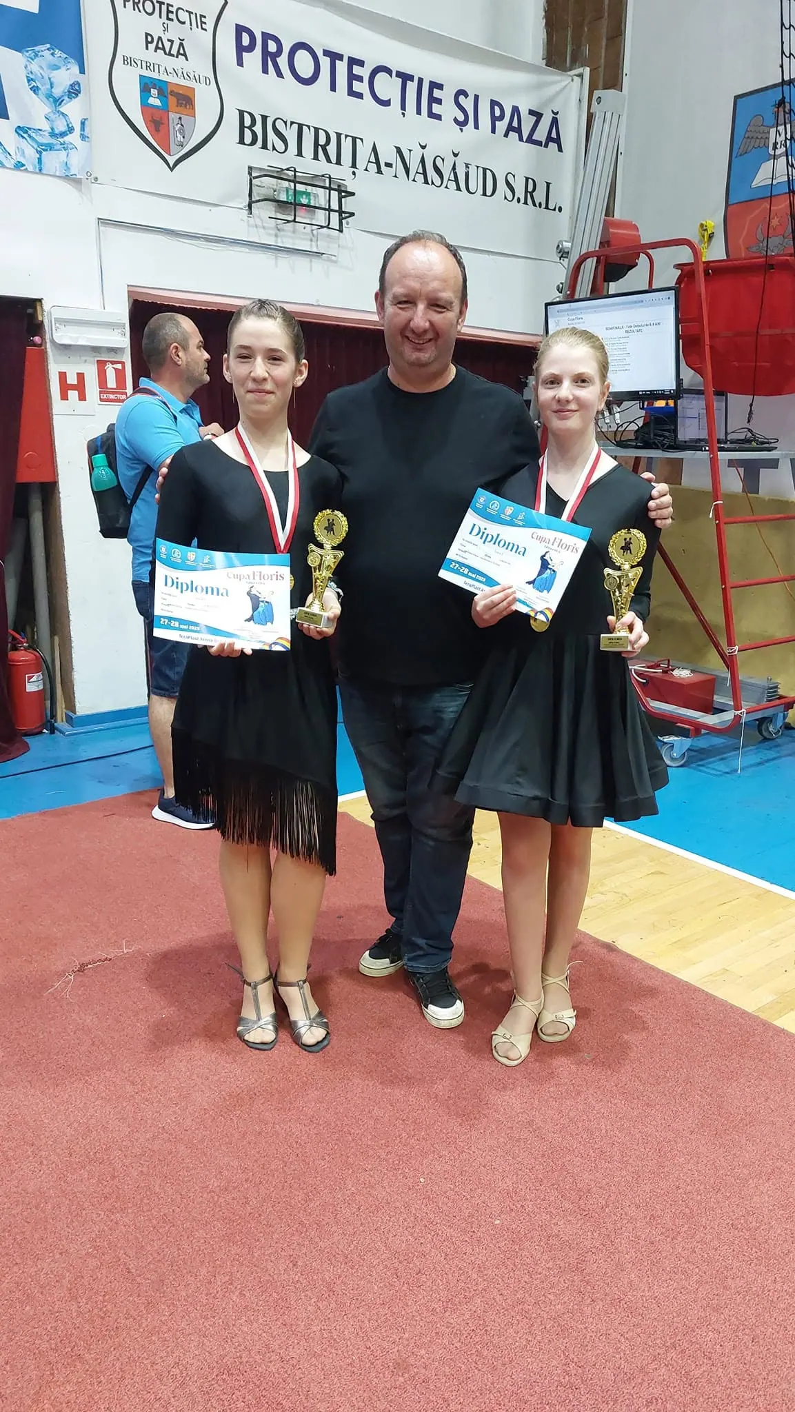 Două dansatoare cu medalii, cupe și diplome Cupa Floris, zâmbind alături de antrenorul lor într-o sală de sport