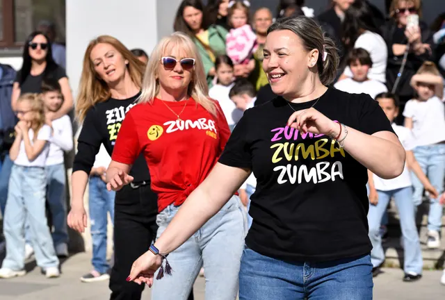 Trei instructoare zâmbind în tricouri Zumba în timpul unei demonstrații în aer liber, în fața publicului